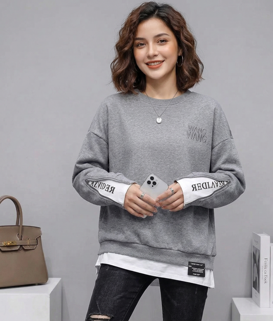 Woman wearing a gray sweatshirt with white sleeves, holding a phone, standing next to a handbag and books on a gray background.